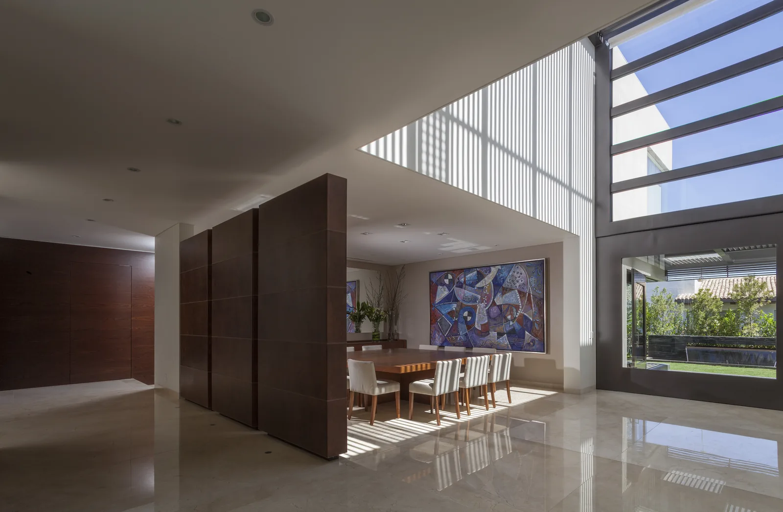 Comedor de doble altura con tragaluz de pérgola, muro de madera y piso de mármol, abierto al jardín.